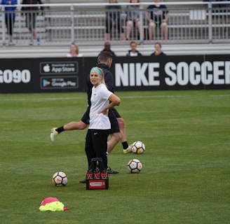 A person in sports attire stands on a soccer field, surrounded by several soccer balls and a carrier with bottles. The field is bordered by advertisements for Nike Soccer and app stores. Several people sit in bleachers in the background.