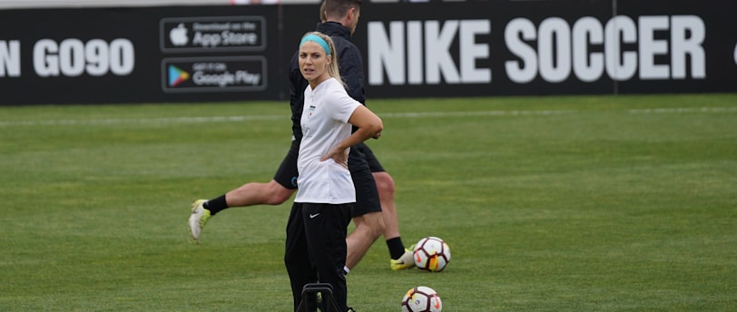 A person in sports attire stands on a soccer field, surrounded by several soccer balls and a carrier with bottles. The field is bordered by advertisements for Nike Soccer and app stores. Several people sit in bleachers in the background.