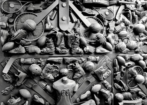 A complex arrangement of various sports equipment and attire, including tennis rackets, soccer balls, basketballs, helmets, shoes, gloves, and a mannequin torso wearing a T-shirt. The items are layered and artistically displayed in a monochrome color scheme, creating an intricate texture and visual pattern.