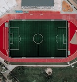 An aerial view of a large sports complex featuring a central green football field surrounded by a red running track. Adjacent to the track, there is a parking area, buildings, and pathways. Nearby, there are additional sports courts in different colors, including a blue swimming pool and orange basketball courts.