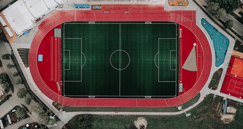 An aerial view of a large sports complex featuring a central green football field surrounded by a red running track. Adjacent to the track, there is a parking area, buildings, and pathways. Nearby, there are additional sports courts in different colors, including a blue swimming pool and orange basketball courts.