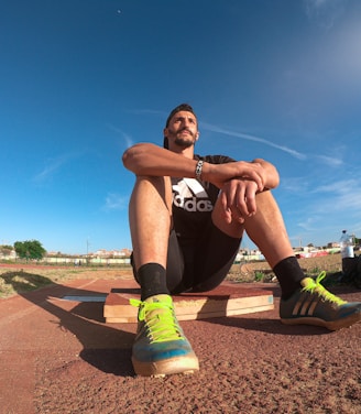 A person wearing a black athletic outfit is seated on a wooden platform in an outdoor sports field. The individual is relaxed, with arms resting on knees. The field is expansive with a clear blue sky and scattered clouds above. A brightly colored running shoe and athletic gear are visible, along with a water bottle and a blue backpack placed beside the person. Lampposts and residential buildings can be seen in the background.