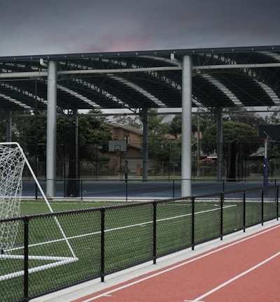 An outdoor sports facility featuring a running track and a soccer goal. The track is red, surrounded by a black metal fence, and the field is covered with artificial green turf. In the background, a basketball court is visible under a covered roof with metal supports. The sky is overcast, adding a moody atmosphere.
