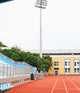 A sports stadium with a red running track, blue seating area, and a large floodlight. The background features a yellow building with blue and white accents, surrounded by green trees. The atmosphere is clean and empty, with clear skies above.