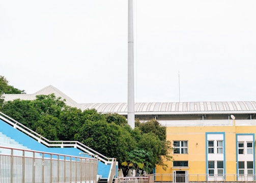 A sports stadium with a red running track, blue seating area, and a large floodlight. The background features a yellow building with blue and white accents, surrounded by green trees. The atmosphere is clean and empty, with clear skies above.