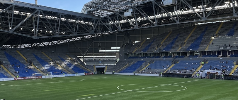 A modern sports stadium with a large, green soccer field and surrounding seating areas. Blue and yellow seats are visible, with some spectators scattered across the stands. The stadium is partially covered with a steel lattice roof, showing a clear blue sky. Several security personnel in high-visibility vests are present.