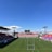 A large sports stadium with red and blue seating overlooks a green field with a goalpost at the center. A few people are scattered across the field, with some tents and a gathering of individuals on the right side. The sky is clear and blue with some clouds visible in the distance. A large screen or scoreboard is situated at the far end of the field.