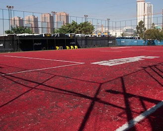 A sports court with a red playing surface and white lines, surrounded by a black mesh fence. In the background, tall buildings are visible, along with some trees and a few yellow chairs positioned near the fence.