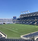 A large, empty sports stadium with blue seats surrounding a well-maintained green grass field. Bright sunlight creates clear shadows, and there are two goal posts on opposite ends of the field. A big electronic scoreboard displays some graphics, and the sky is clear and blue.
