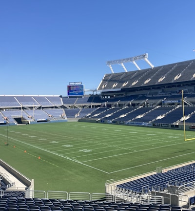 A large, empty sports stadium with blue seats surrounding a well-maintained green grass field. Bright sunlight creates clear shadows, and there are two goal posts on opposite ends of the field. A big electronic scoreboard displays some graphics, and the sky is clear and blue.