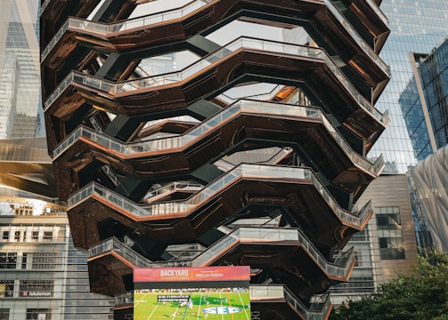 A large, spiral architectural structure with multiple levels stands prominently in an urban setting. In front of the structure, a large screen displays a sports broadcast, and several people are gathered around, some sitting and others standing. The scene is set outdoors, with towering modern buildings surrounding the area. The ground is paved with a geometric pattern of gray and white tiles.